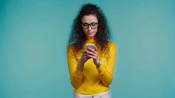 Jovem mulher em óculos usando smartphone isolado em azul — Fotografia de Stock