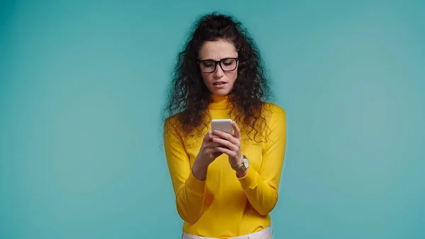 Unzufriedene junge Frau mit Brille textet auf Smartphone isoliert auf blauem Grund — Stockfoto