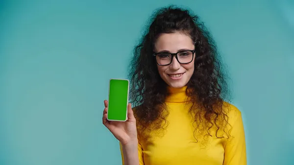 Allegra giovane donna in occhiali che tiene smartphone con schermo verde isolato su blu — Foto stock