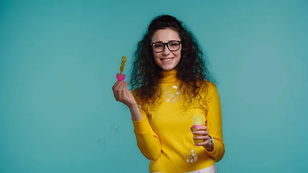 Joyful young woman in glasses holding bottle with soap bubbles isolated on blue — Stock Photo