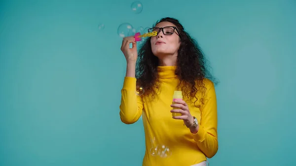 Femme brune dans des lunettes soufflant des bulles de savon isolé sur bleu — Photo de stock