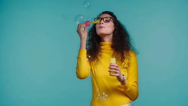 Brunette woman in glasses blowing soap bubbles isolated on blue — Stock Photo