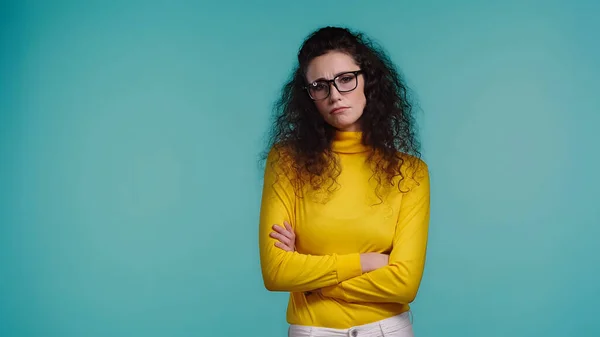 Unzufriedene junge Frau mit Brille und Rollkragen, die mit verschränkten Armen auf blauem Grund steht — Stockfoto