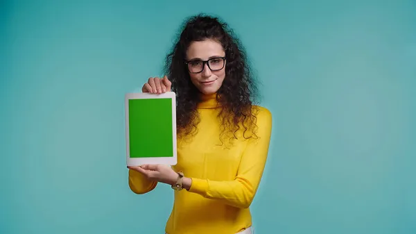 Молодая веселая женщина в очках, держа цифровой планшет с зеленым экраном изолированы на синий — стоковое фото