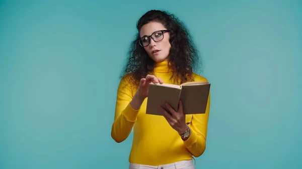 Скучающая женщина в очках, читающая книгу, изолированную на синий — стоковое фото