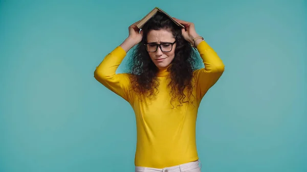 Upset young woman in glasses holding book above head isolated on blue — Stock Photo