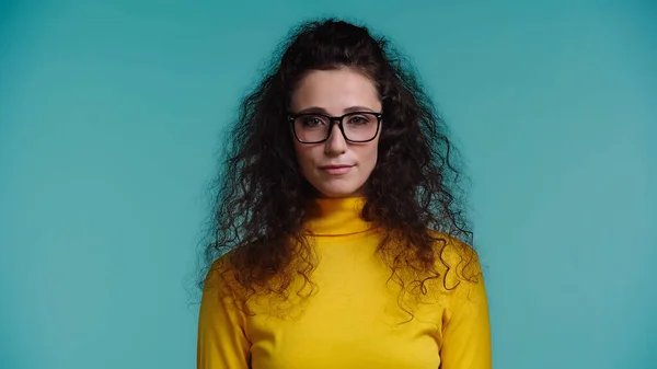 Молода жінка в жовтому горлику і окулярах дивиться на камеру ізольовано на синьому — стокове фото