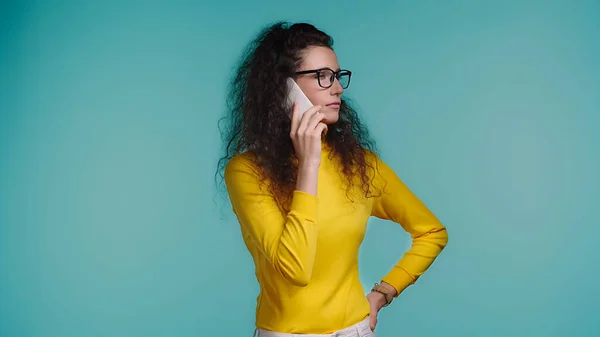 Молодая женщина в очках разговаривает на смартфоне изолированы на синий — стоковое фото