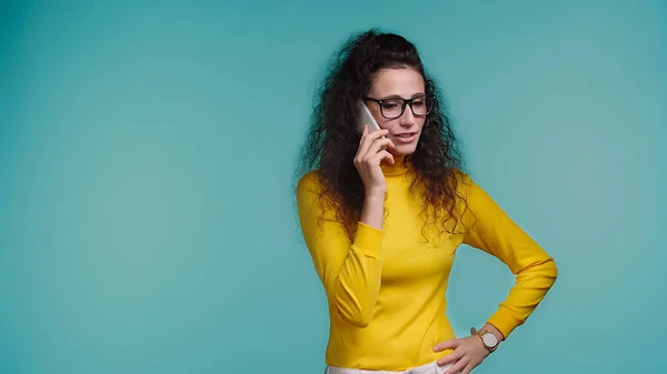 Young woman in glasses talking on smartphone while standing with hand on hip isolated on blue — Stock Photo