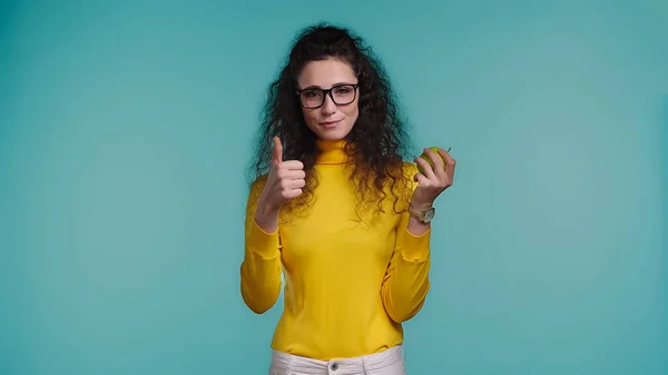 Young curly woman holding apple while showing thumb up isolated on blue — Stock Photo