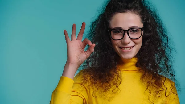 Радісна молода жінка в окулярах демонструє знак ОК ізольовано на синьому — стокове фото