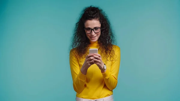 Улыбающаяся молодая женщина в очках с помощью мобильного телефона изолированы на синий — стоковое фото