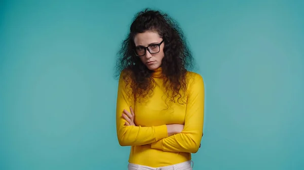 Offended young woman in glasses and turtleneck standing with crossed arms isolated on blue — Stock Photo