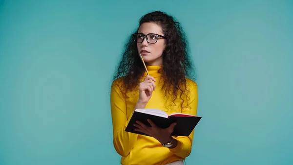 Mujer joven pensativa en gafas y cuello alto sosteniendo portátil y pluma aislada en azul - foto de stock