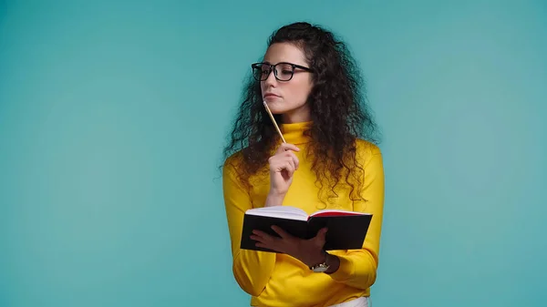 Nachdenkliche junge Frau mit Brille und Rollkragen, Notizbuch und Stift isoliert auf blauem Grund — Stockfoto