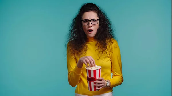 Schockierte junge Frau mit Popcorn-Eimer auf blauem Grund — Stockfoto