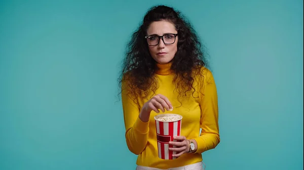 Curly young woman watching movie and holding popcorn bucket isolated on blue — Stock Photo
