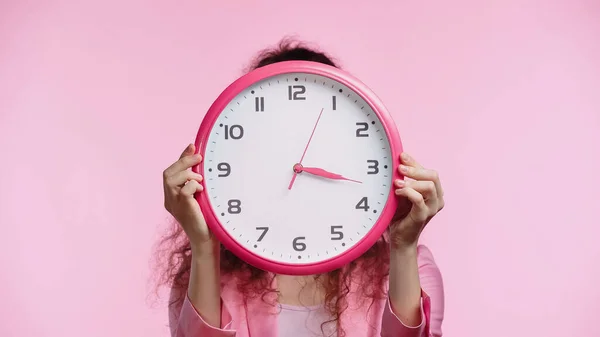 Молода жінка затемнює обличчя з настінним годинником ізольовано на рожевому — стокове фото