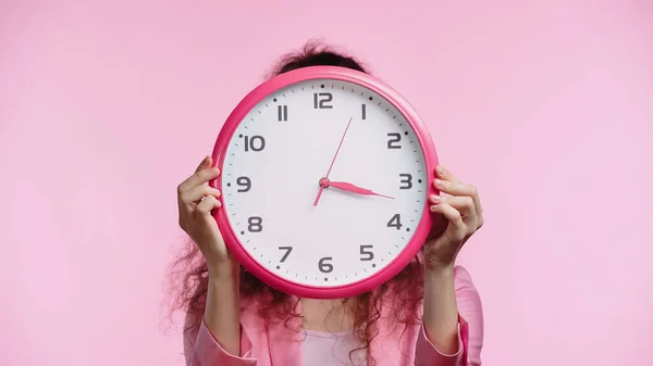 Junge Frau verdeckt Gesicht mit Wanduhr isoliert auf rosa — Stockfoto