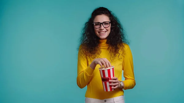 Alegre joven mujer sosteniendo palomitas de maíz cubo mientras viendo película aislado en azul - foto de stock