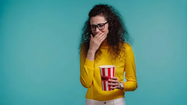 Glückliche junge Frau hält Popcorn-Eimer in der Hand und schaut Komödienfilm isoliert auf blauem Grund — Stockfoto