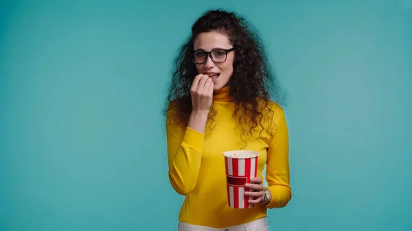 Curly young woman eating popcorn while watching movie isolated on blue — Stock Photo