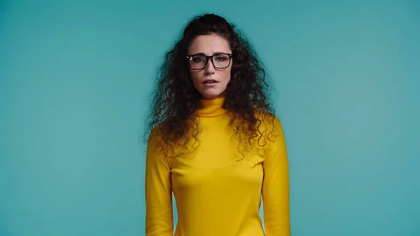 Brünette und lockige junge Frau mit Brille, die isoliert auf blauem Grund in die Kamera blickt — Stockfoto