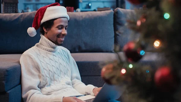 Homme afro-américain joyeux chapeau santa en utilisant un ordinateur portable près de l'arbre de Noël flou — Photo de stock