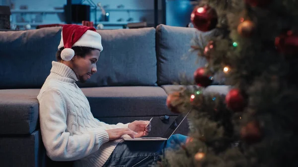 Side view of african american freelancer in santa hat using laptop near blurred christmas tree — Stock Photo