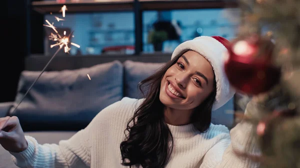 Happy african american woman in santa hat holding sparkler near blurred christmas tree — Stock Photo