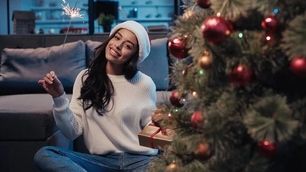 Feliz africana americana mujer en santa hat celebración sparkler y presente cerca borrosa navidad árbol - foto de stock