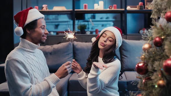 Cheerful african american man in santa hat and woman blowing kiss while holding sparkler near christmas tree — Stock Photo