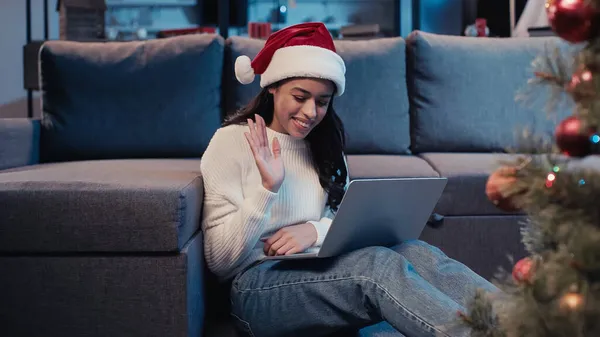 Happy african american woman in santa hat waving hand while having video call — Stock Photo