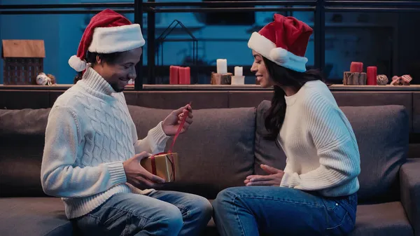 African american man holding christmas present near happy woman in santa hat — Stock Photo