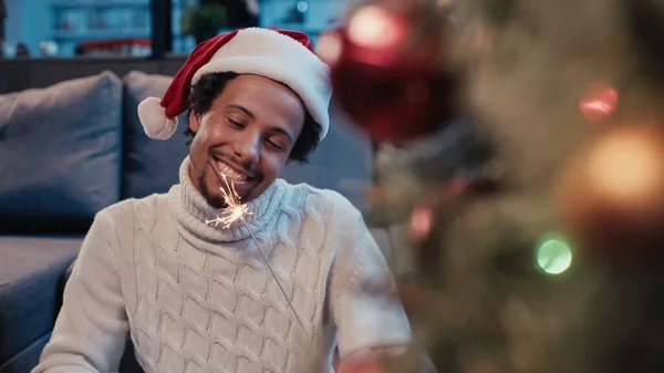 African american man looking at sparkler near blurred christmas tree — Stock Photo