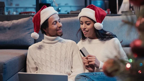 Fröhliches afrikanisch-amerikanisches Paar mit Weihnachtsmützen beim Online-Shopping in der Nähe des verschwommenen Weihnachtsbaums — Stockfoto