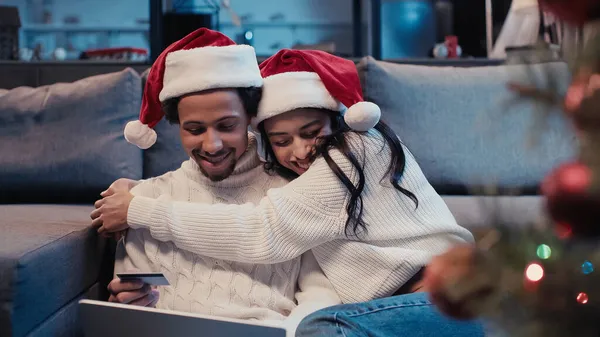 Happy african american couple in santa hats hugging while online shopping near blurred christmas tree — Stock Photo