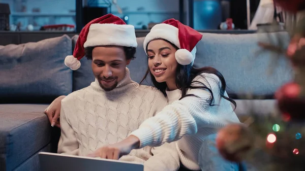 Glückliches afrikanisch-amerikanisches Paar in Weihnachtsmannmützen mit Laptop in der Nähe des verschwommenen Weihnachtsbaums — Stockfoto