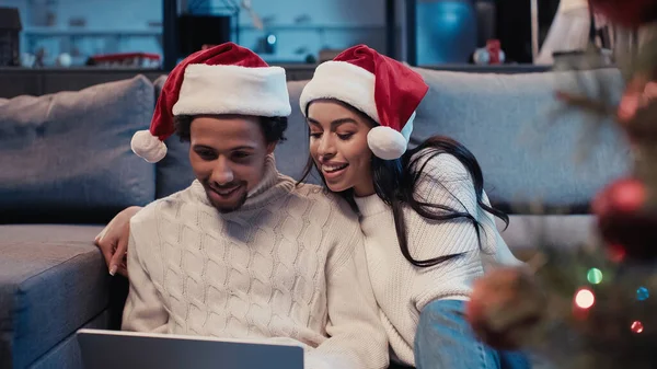 Cheerful african american couple in santa hats using laptop near blurred christmas tree — Stock Photo