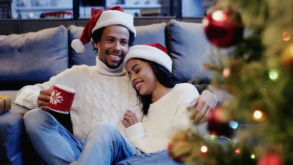 Allegro uomo africano americano che tiene la tazza e sorride con la ragazza vicino all'albero di Natale — Foto stock