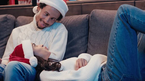 African american man in santa hat sticking out tongue near woman on cough — Stock Photo