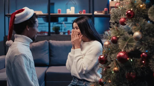 Alegre africano americano mujer riendo mientras mirando novio en santa hat cerca de árbol de Navidad - foto de stock