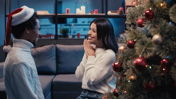 Alegre africana americana mujer sonriendo mientras mira novio en santa hat cerca de árbol de navidad - foto de stock