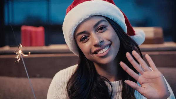 Happy african american woman in santa hat waving hand and holding sparkler — Stock Photo