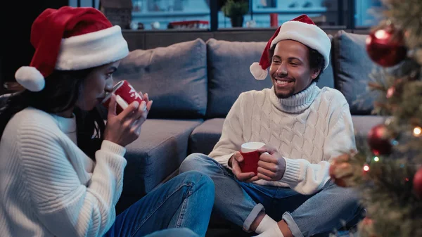 Happy african american man in santa hat holding cup and looking at girlfriend drinking near blurred christmas tree — Stock Photo