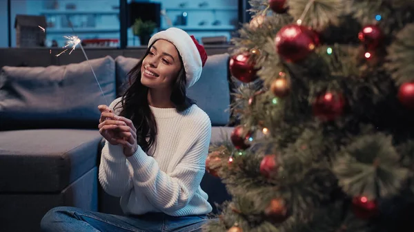 Feliz africana americana mujer en santa hat holding sparkler cerca borrosa decorado abeto - foto de stock