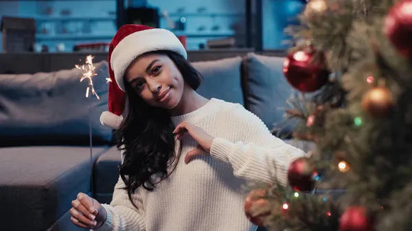 Happy african american woman in santa hat holding sparkler and gesturing near blurred pine tree — Stock Photo