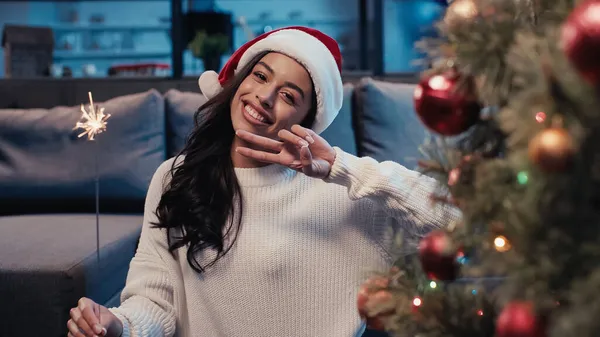 Happy african american woman in santa hat holding sparkler showing peace sign near blurred fir — Stock Photo