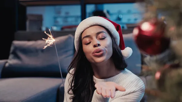 Young african american woman in santa hat holding sparkler and sending air kiss near blurred fir — Stock Photo