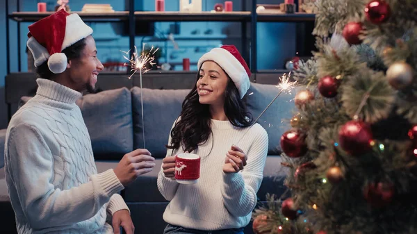 Happy african american couple in santa hats holding cup and sparklers near blurred christmas tree — Stock Photo